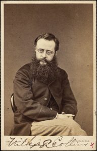 Black and white photograph of Wilkie Collins: a man with short hair, glasses and a bushy beard.