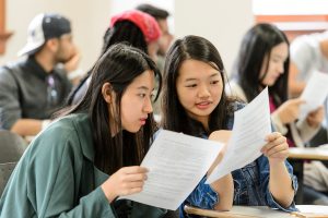 Two students review an academic writing assignment together. More students in the class are present in the background.