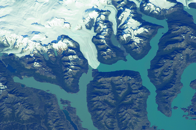 A photograph shows an aerial view of a land mass. The white mass of a glacier is shown near the top left quadrant of the photo and leads to two branching blue rivers. The open land is shown in brown.