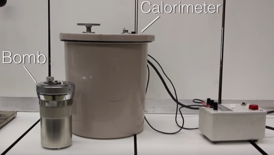 A stainless steel cylinder with a screw cap is labeled "bomb". Next to it is a larger brown plastic tub labeled calorimeter. Connected to both is an electrical control box.