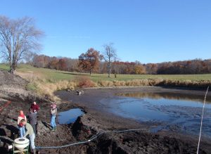 A pond in a valley next to a hill. Some people are working in the lower left corner, where dark colored organic soil is visible