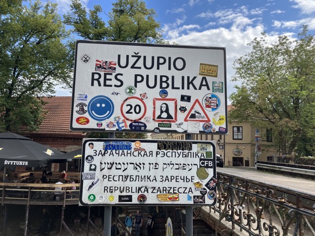 sign that says: Užupio Res publika, Республика заречье, names of the place in several other languages, and lots of stickers on the sign