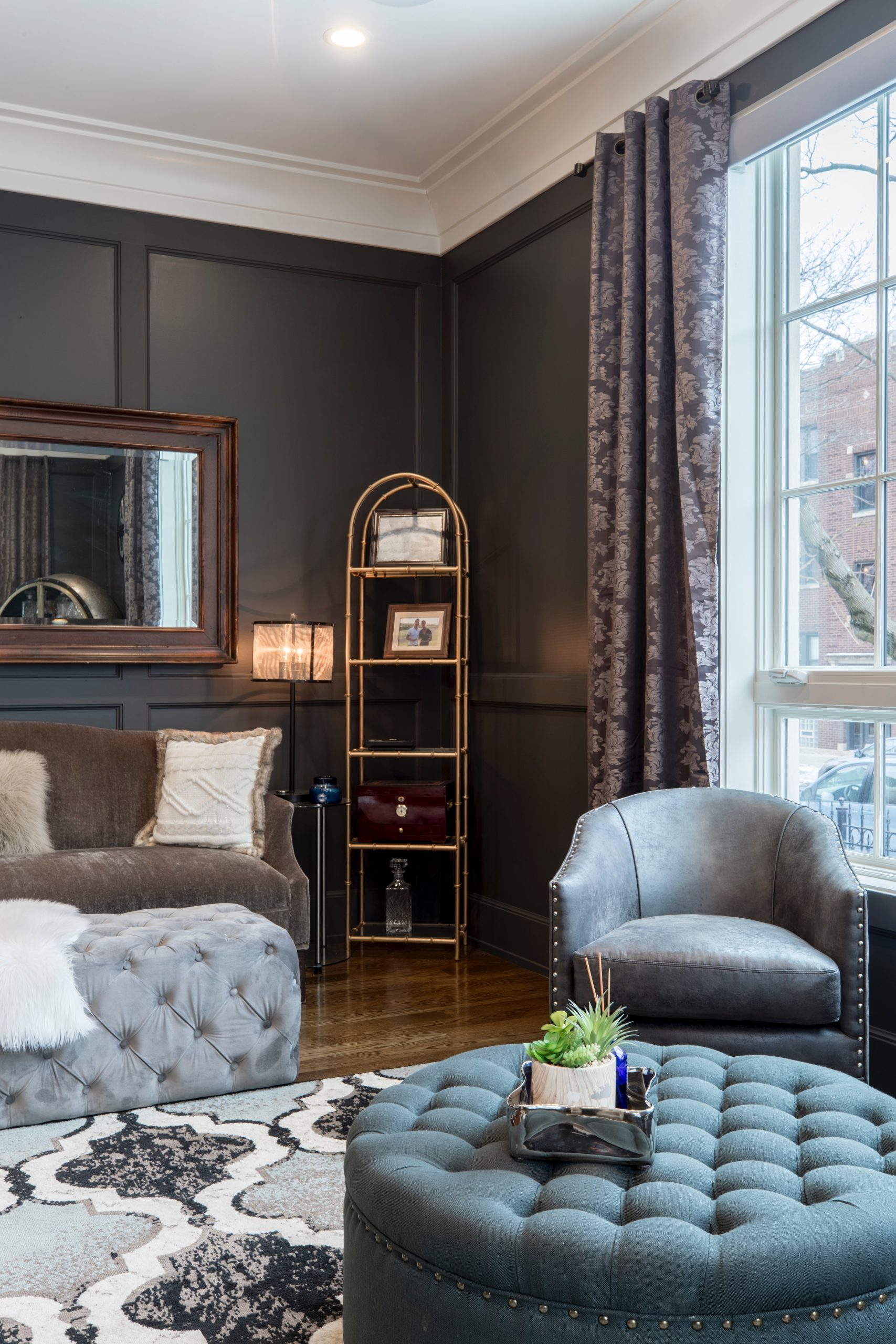 Bright living room with forest green paneled walls, a grey, white, and black rug, a green ottoman, a grey ottoman, a gray leather chair, a light brown fabric couch, a large mirror, a golden shelf with pictures and trinkets, and dark curtains