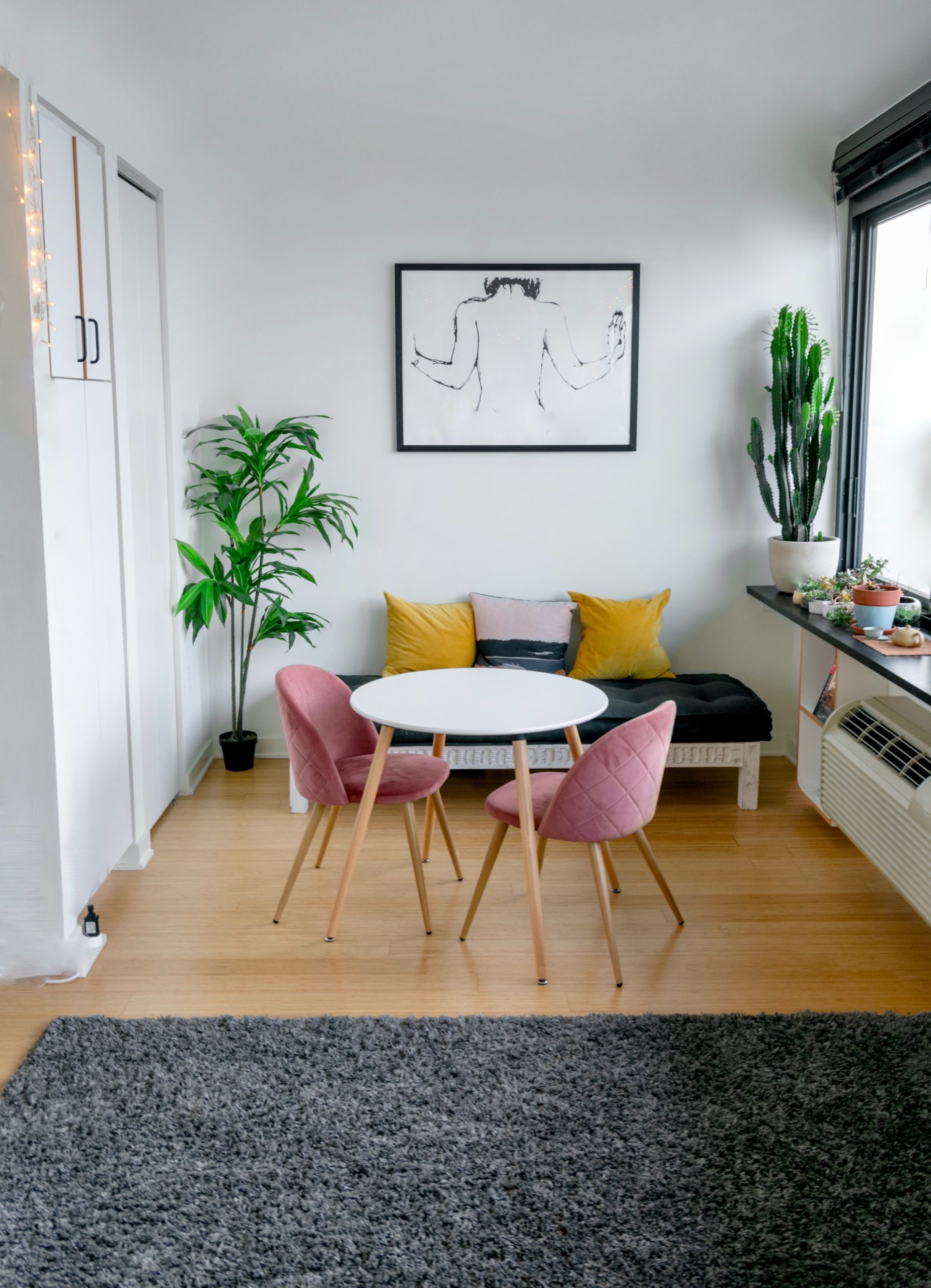 Living room area with a small white table, two pink chairs, a black and white couch with yellow and white pillows, white walls, bright natural light, a cactus plant, a large house plant, a rough sketch on the wall, and a fuzzy grey rug