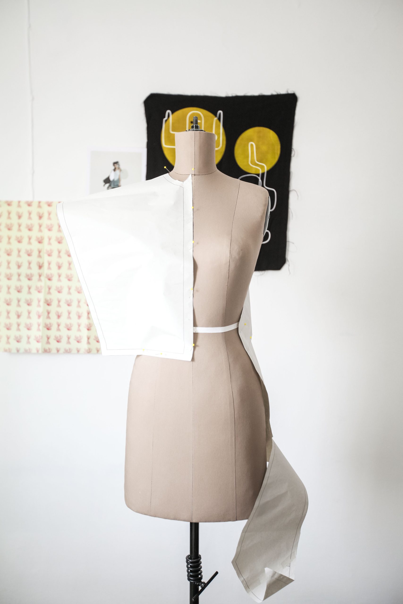 Mannequin with fabric piece pinned to it, in front of a white wall decorated wiith fabric squares