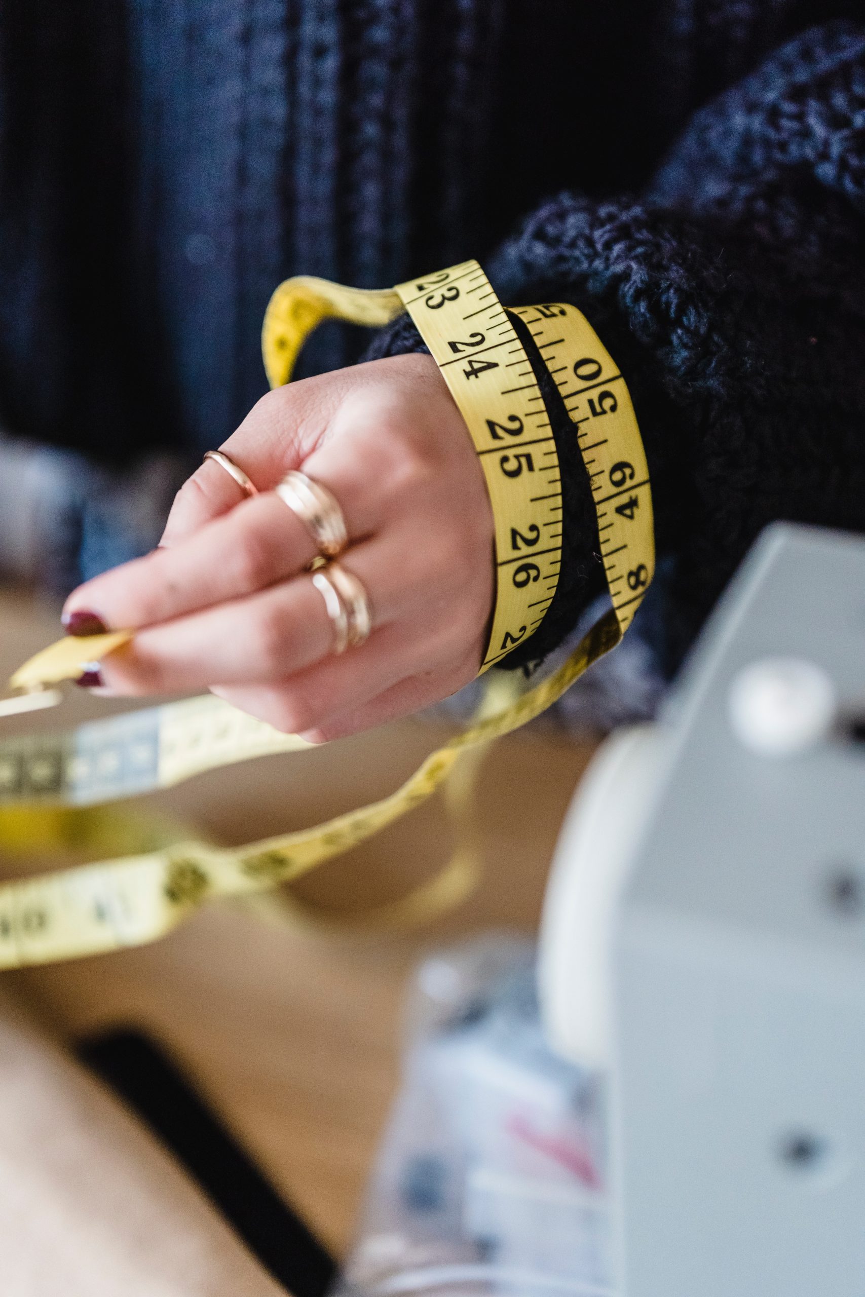 Hand adorned with gold rings wrapped in a measuring tape with a sewing machine in the background