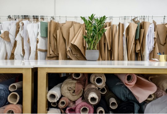 Rolls of fabric rest on a lower shelf, and garment pieces hand on a clothing rack